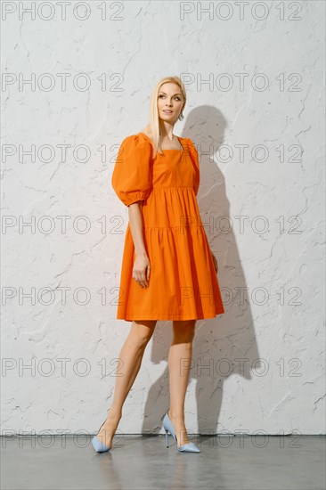 Stylish adult woman in overknee orange sundress posing near white textured wall