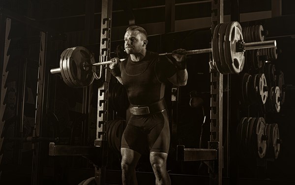 The weightlifter stands with a very heavy barbell on his shoulders. He is at the top and is preparing to sit down