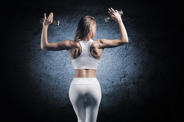 Adorable girl is training in the gym. She lifts the dumbbells up. Back view. The concept of bodybuilding