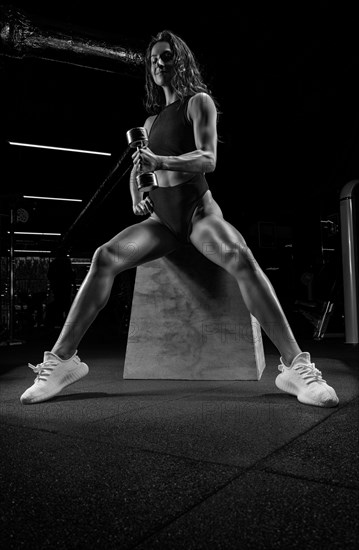 Tall athletic woman posing in the gym on a bench with dumbbells. Biceps pumping. Fitness and bodybuilding concept.