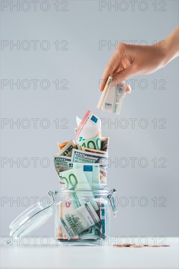 Female hand puts a bill into a glass piggy bank full of money. Thrift concept.