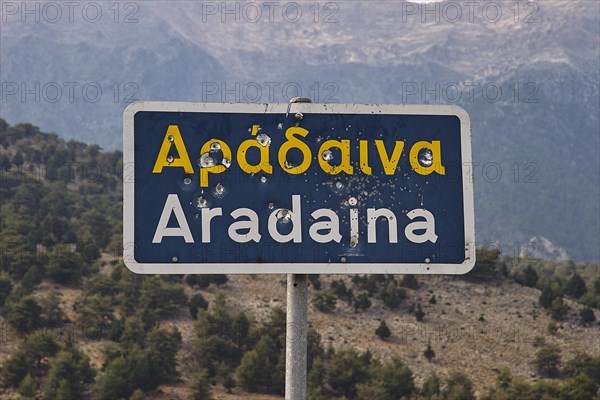 Damaged road sign with Greek and Latin lettering in front of a mountain landscape, Aradena Gorge,