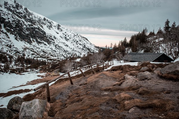 A serene winter landscape with a cabin, snow-covered mountain, and wooden fence at dusk. Poland,