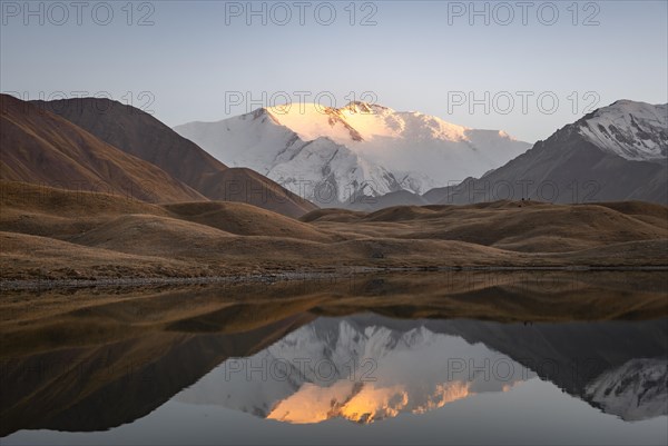 White glaciated and snowy mountain peak Pik Lenin at sunset, mountains reflected in a lake between