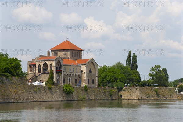 A historic building stands by a quiet river, surrounded by green nature and a light cloudy sky,