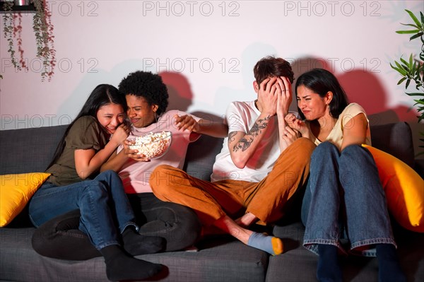 Multi-ethnic friends on a terror movie night at home sitting on the sofa eating popcorn and