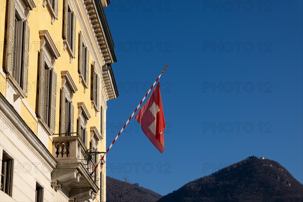 Swiss Flag on City Hall and Mountain Peak Monte Bre Against Blue Clear Sky in a Sunny Day in