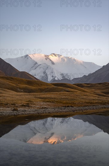 White glaciated and snowy mountain peak Pik Lenin at sunset, mountains reflected in a lake between