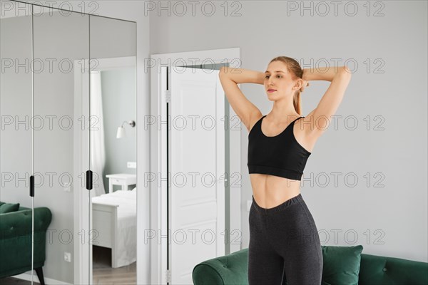 Young sporty woman warming up in her bright apartment doing turns and bends with her arms behind