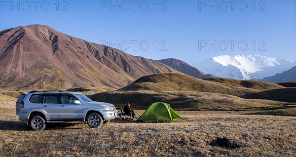 An off-road vehicle stands next to a pitched tent in a mountain landscape with snow-capped peaks in