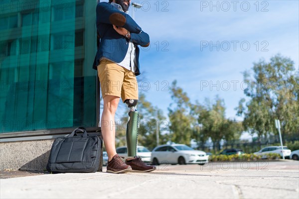Businessman with prosthetic leg waiting patiently leaning on a building with arms crossed in the