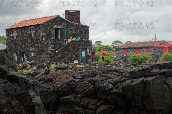 People standing next to a stone house on lava rock under an overcast sky, North Coast, Santa Luzia,