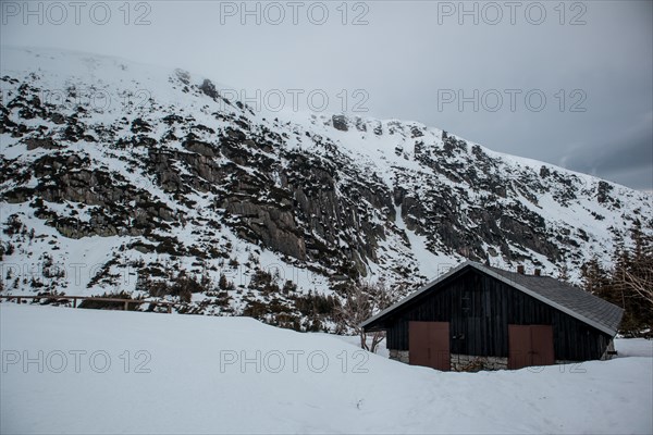 A solitary cabin sits beneath a rugged, snowy mountain landscape under an overcast sky. Poland,