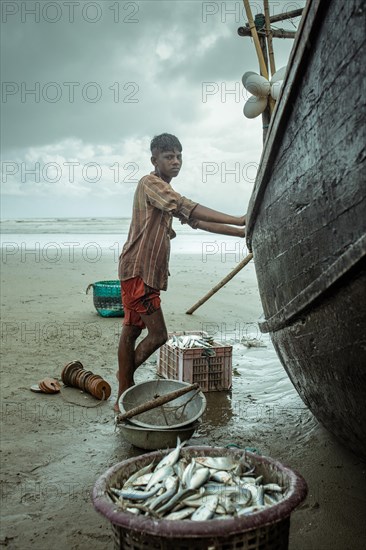 Fisherman by his boat on the beach with the catch of the day during a monsoon shower, Cox's Bazar,