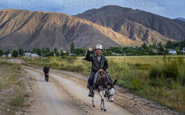 Man in traditional Kyrgyz dress riding on a donkey along a village street and greeting friendly,