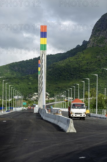A highway with Mauritian emblem on pillar and a truck driving, surrounded by mountains and an