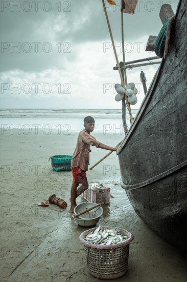 Fisherman by his boat on the beach with the catch of the day during a monsoon shower, Cox's Bazar,
