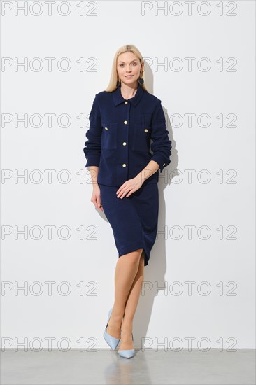 Elegant woman in navy blue button down jacket, skirt and blue heels posing against a white wall