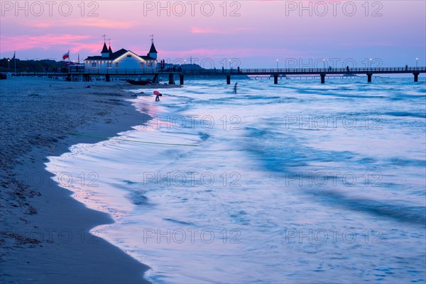 Calm evening mood on the beach with calm waves and two people near a pier with a building at dusk,