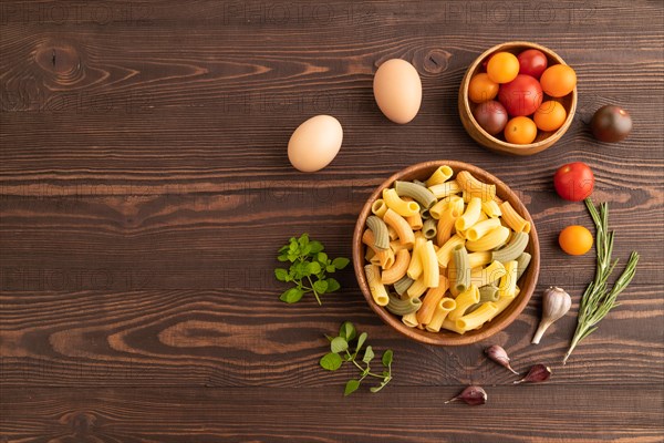 Rigatoni colored raw pasta with tomato, eggs, spices, herbs on brown wooden background. Top view,