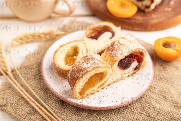 Homemade sweet bun with apricot jam and cup of coffee on white wooden background and linen textile.