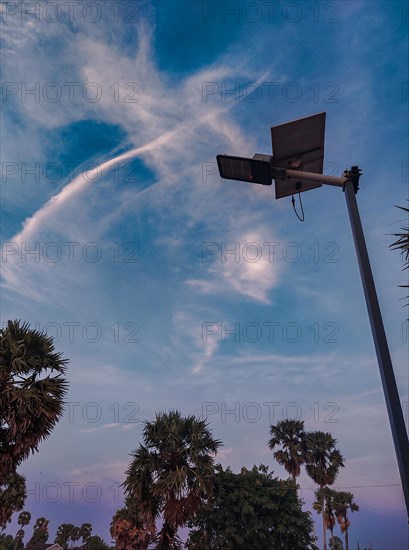 A silhouette of a solar-powered streetlamp against evening sky with clouds and palm trees,