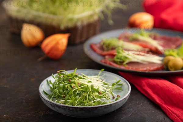 Slices of smoked salted meat with cilantro microgreen on black concrete background and red textile.