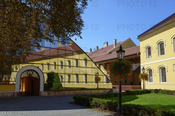A yellow building reflected in a large glass facade, flanked by trees and a paved path, Officers'