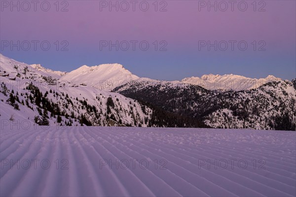 Snow-covered mountains at dusk with a purple sky and a freshly groomed ski slope in the foreground,