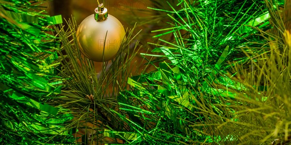 Closeup of gold Christmas ornament and green tensile garland hanging on pine tree in local park in