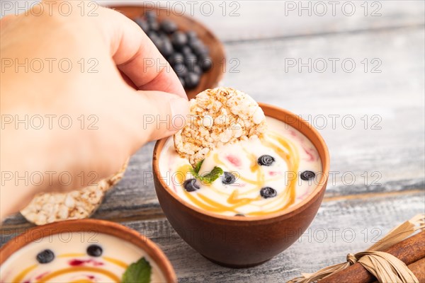 Yoghurt with bilberry and caramel in clay bowl on gray wooden, hand dipping cookies in yogurt, side