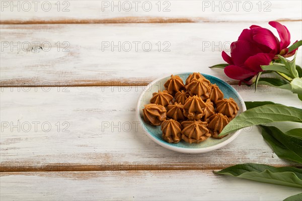 Homemade soft caramel fudge candies on blue plate on white wooden background, peony flower