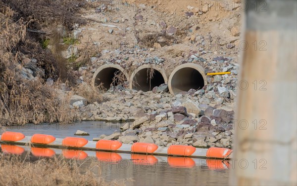 Three water drainage culverts in side of hill that empty into shallow river. Bridge support column