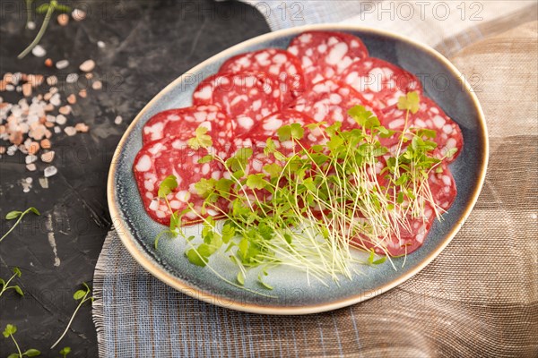 Slices of smoked cervelat salami sausage with spinach microgreen, salt and pepper on black concrete