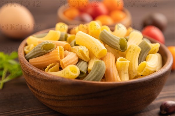 Rigatoni colored raw pasta with tomato, eggs, spices, herbs on brown wooden background. Side view,