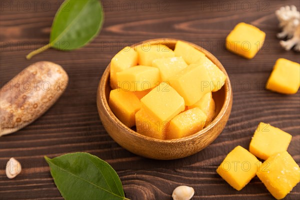 Dried and candied mango cubes in wooden bowls on brown wooden textured background. Side view, close