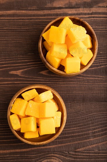 Dried and candied mango cubes in wooden bowls on brown wooden textured background. Top view, flat