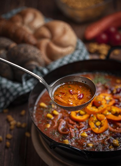 Soup ladle scooping bean soup with sweetcorn and peppers from a pot, basket with pastries in the