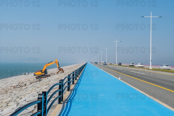 Backhoe sitting on ocean causeway next to blue concrete sidewalk on divided highway with speeding