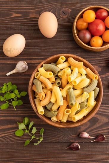 Rigatoni colored raw pasta with tomato, eggs, spices, herbs on brown wooden background. Top view,