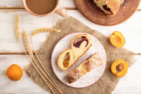 Homemade sweet bun with apricot jam and cup of coffee on white wooden background and linen textile.