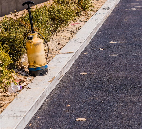 Plastic industrial spray bottle sitting on the curb of parking lot with black asphalt paving and a