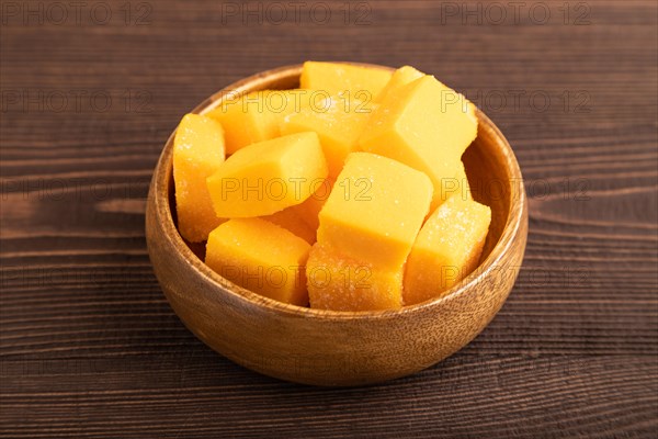 Dried and candied mango cubes in wooden bowls on brown wooden textured background. Side view, close