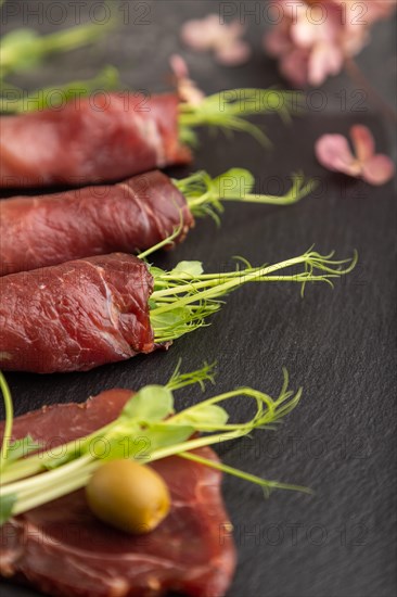 Slices of smoked salted meat with green pea microgreen on black concrete background. Side view,