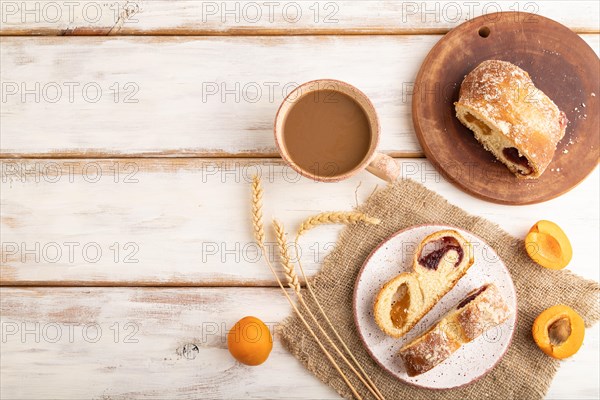 Homemade sweet bun with apricot jam and cup of coffee on white wooden background and linen textile.