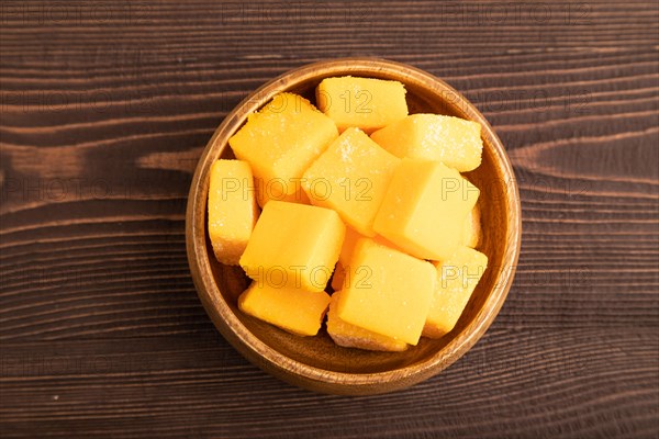 Dried and candied mango cubes in wooden bowls on brown wooden textured background. Top view, flat