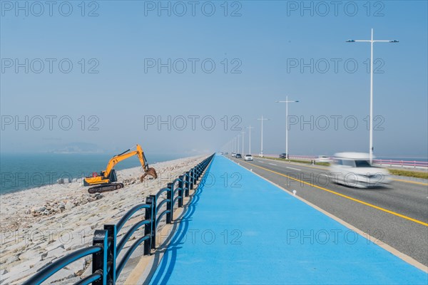 Backhoe sitting on ocean causeway next to blue concrete sidewalk on divided highway with speeding