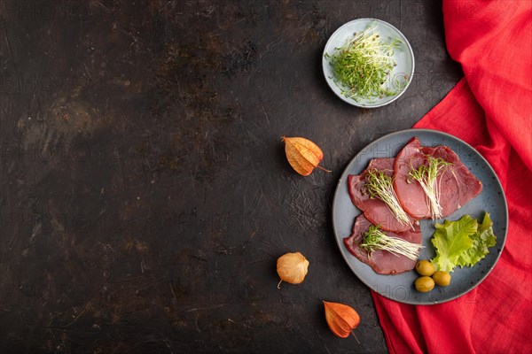 Slices of smoked salted meat with cilantro microgreen on black concrete background and red textile.