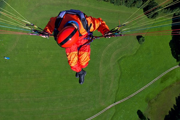 A man in signal red overalls hangs from a paraglider high above the meadows near Lenggries, pilot's