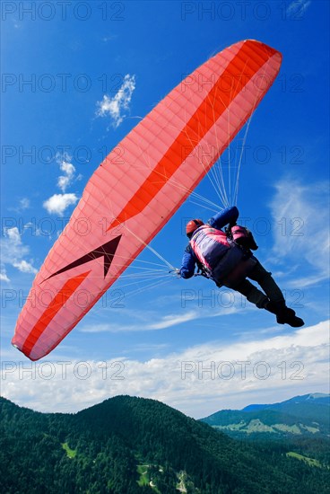 A man hangs in a red and white paraglider high above the meadows near Lenggries, pilot's